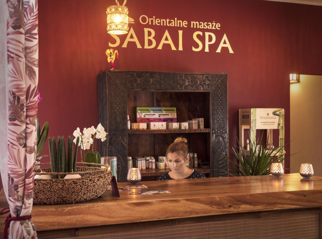 Recepcja w orientalnym SPA z drewnianym blatem, dekoracjami roślinnymi i uśmiechniętą recepcjonistką za ladą, widoczne logo Sabai SPA na ścianie.