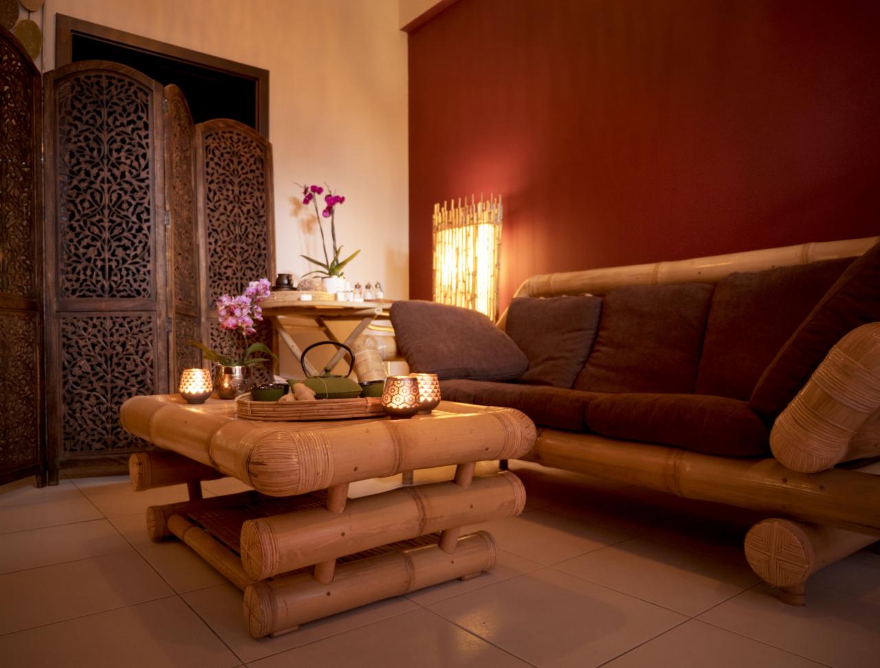 Stylowe wnętrze z bambusowymi meblami: sofa z brązowymi poduszkami, stolik z herbatą i lampionami, ozdobny parawan i kwiaty, tworzące relaksującą atmosferę.