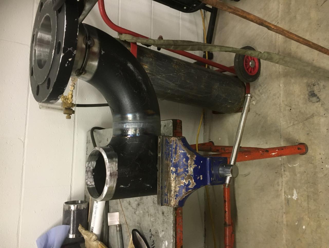 Zespawany element rury z kołnierzem mocowany w starym imadle; widoczny wózek transportowy z butlą gazową w tle.