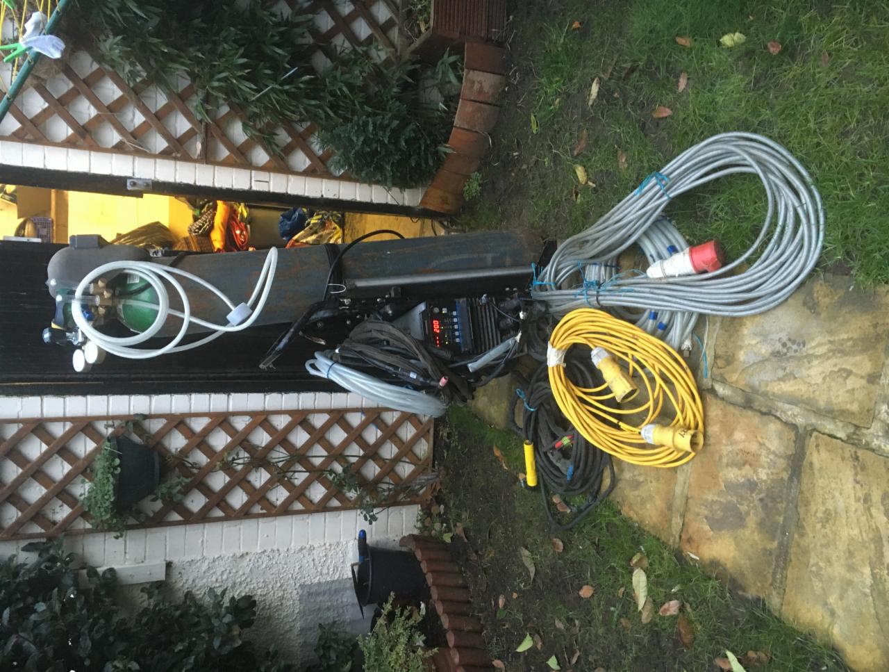 Sprzęt do obróbki metali z butlą gazową, przewodami i urządzeniem sterującym leżący na zewnątrz budynku, na trawie i kamiennej nawierzchni.