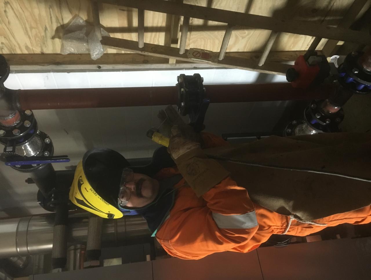 Pracownik w kombinezonie ochronnym i masce spawalniczej wykonuje prace przy rurociągu z licznymi kołnierzami, widoczne elementy instalacji w pomieszczeniu.