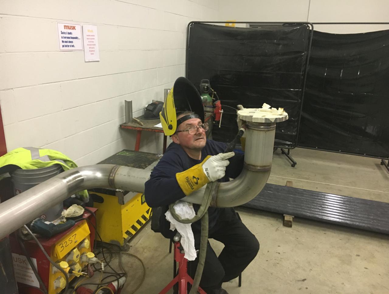 Pracownik w okularach i z podniesioną przyłbicą spawalniczą, w żółtych rękawicach, wskazujący na spawany element rury z kołnierzem, w tle czarna kurtyna spawalnicza i sprzęt spawalniczy.