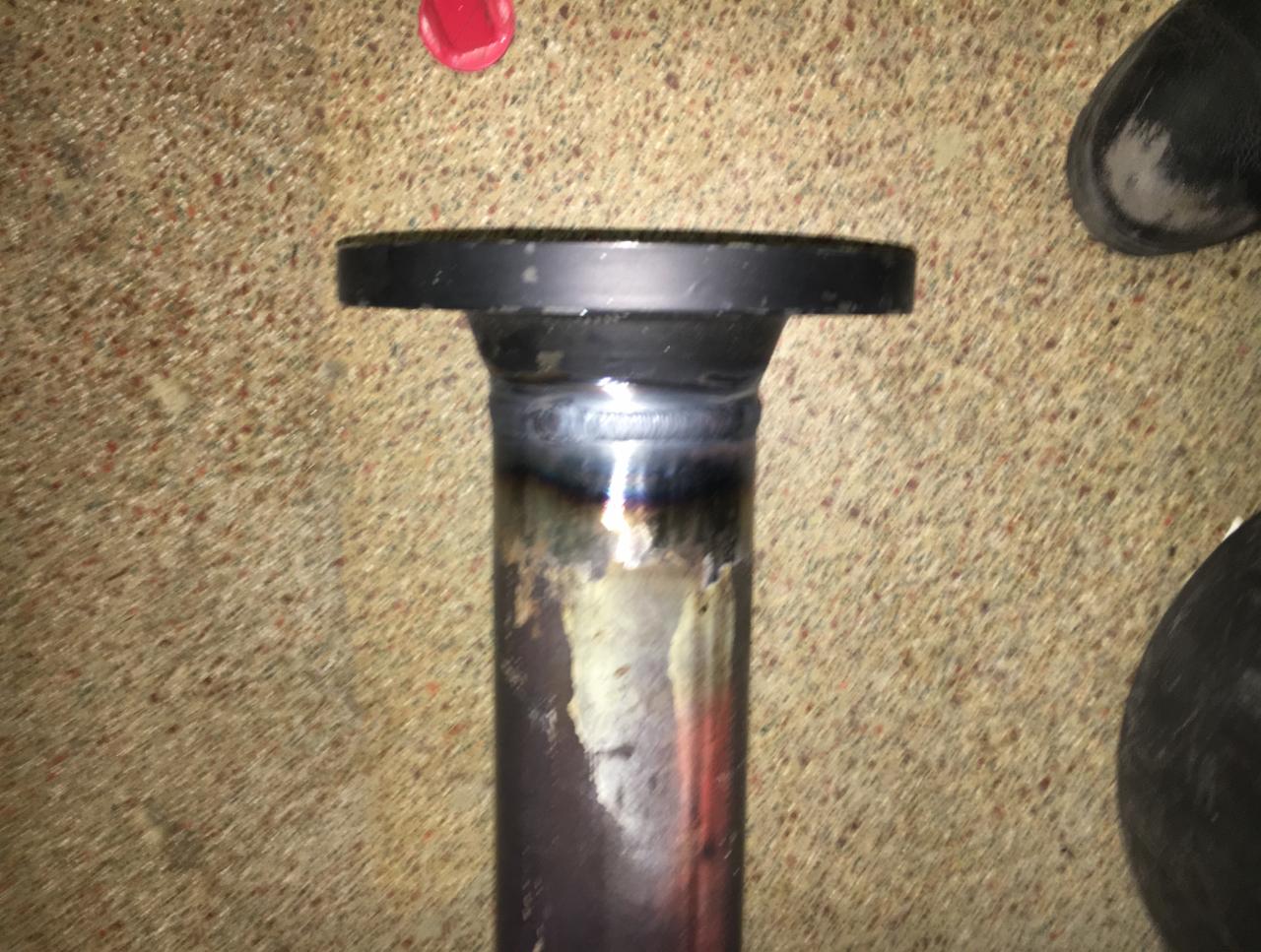 Spoina na rurze stalowej z przymocowanym kołnierzem, widoczne ślady ciepła i odbarwienia metalu, ujęcie z góry na tle podłogi z lastryko.