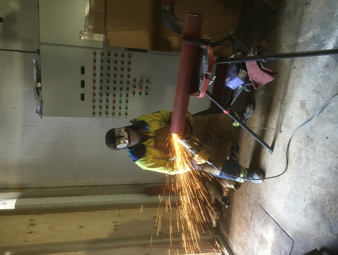 Pracownik w kombinezonie ochronnym i masce szlifierskiej obrabia rurę szlifierką kątową, iskry rozchodzą się promieniście, w tle widoczna szafa sterownicza i fragment podłogi warsztatu.