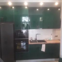 Nowoczesna kuchnia z zielonymi, lakierowanymi frontami szafek górnych, czarnym piekarnikiem i lodówką side-by-side; drewniany blat roboczy z czarnym zlewem i baterią.