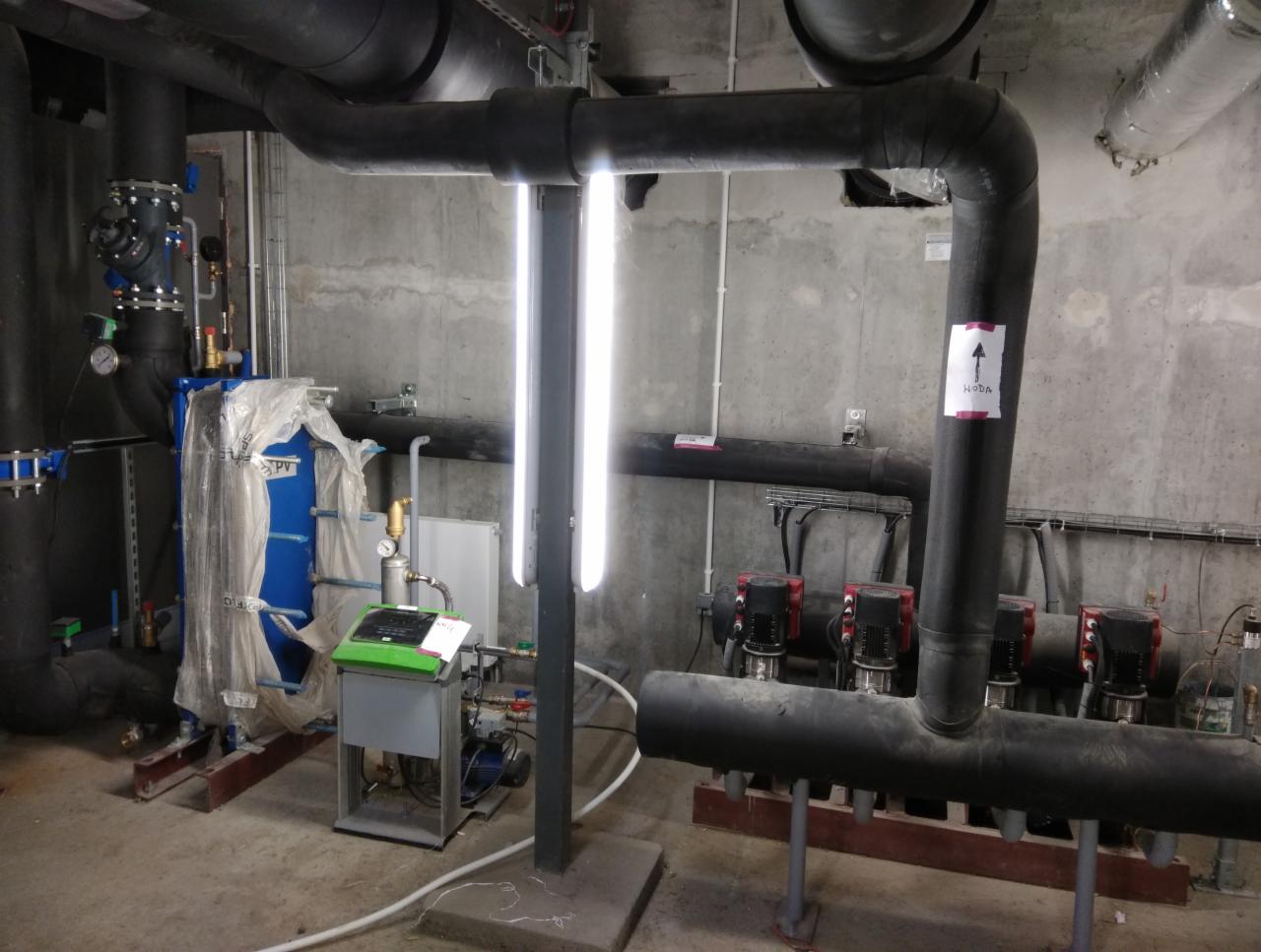 Węzeł cieplny w surowym pomieszczeniu z betonowymi ścianami, widoczne rury, pompy, filtry i aparatura kontrolna, oświetlone dwiema lampami jarzeniowymi.