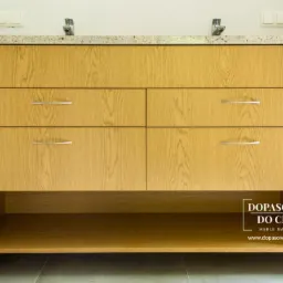 Jasna, dębowa szafka pod umywalkę z czterema szufladami i otwartą półką, zwieńczona blatem z jasnego granitu, z widocznym logo firmy na dole po prawej stronie.