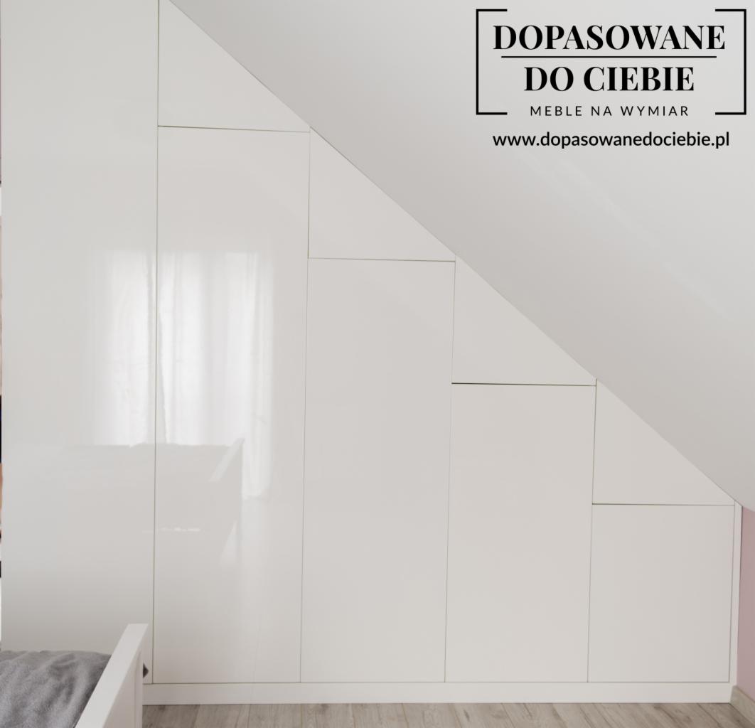 Biała, lakierowana zabudowa wnękowa pod skosem, dopasowana do kształtu poddasza, widoczny fragment łóżka, na górze logo firmy.
