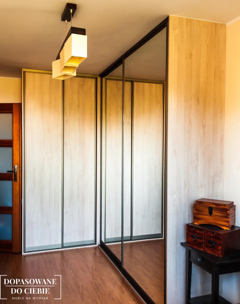 Wnętrze z dużą szafą przesuwną z lustrzanymi drzwiami i drewnopodobnym wykończeniem, uzupełnione dekoracyjną komodą z drewna egzotycznego i nowoczesną lampą sufitową.