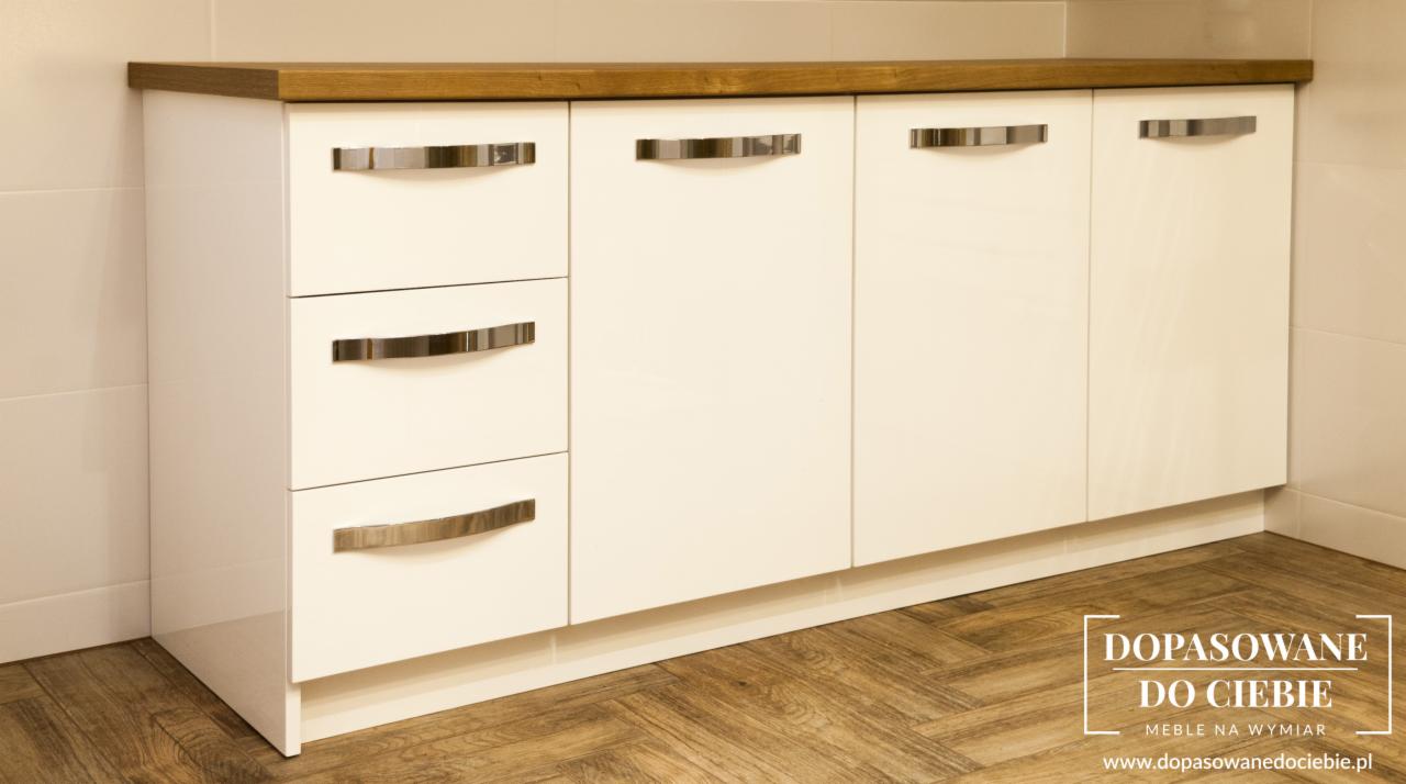 Białe szafki kuchenne z drewnianym blatem i chromowanymi uchwytami, widoczne na tle płytek i parkietu ułożonego w jodełkę, z naklejonym logo firmy na parkiecie.