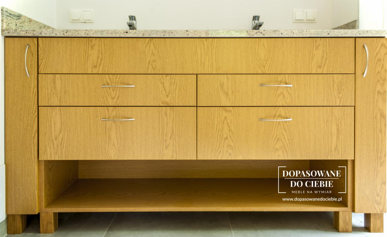 Jasna, dębowa szafka pod umywalkę z czterema szufladami i otwartą półką, zwieńczona blatem z jasnego granitu, z widocznym logo firmy na dole po prawej stronie.