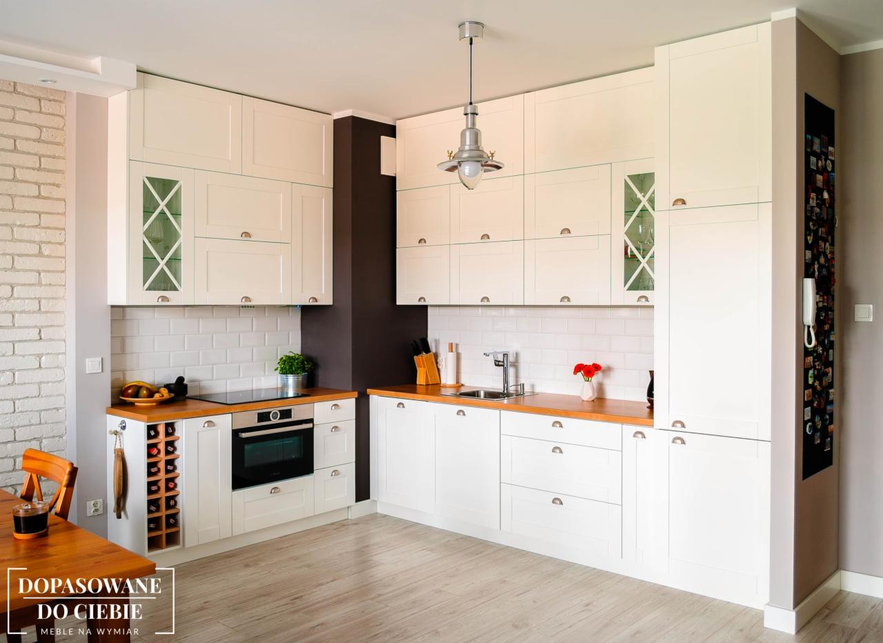 Jasna kuchnia z białymi frontami, drewnianymi blatami i dekoracyjnymi szprosami w szafkach wiszących, oświetlona wiszącą lampą w stylu industrialnym.