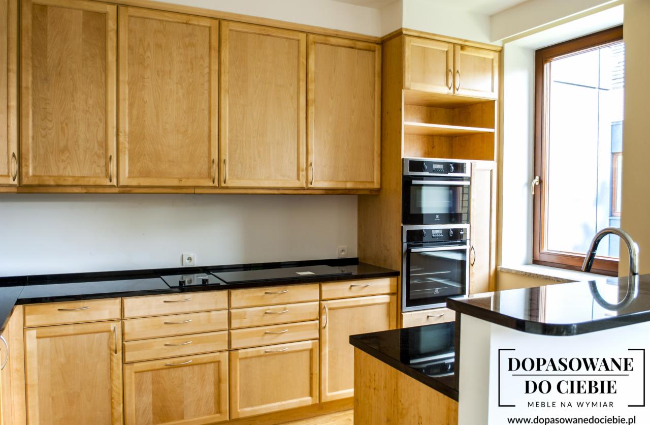 Jasna, drewniana zabudowa kuchenna z czarnym blatem, widoczne dwie wbudowane piekarniki oraz okno z chromowaną baterią.
