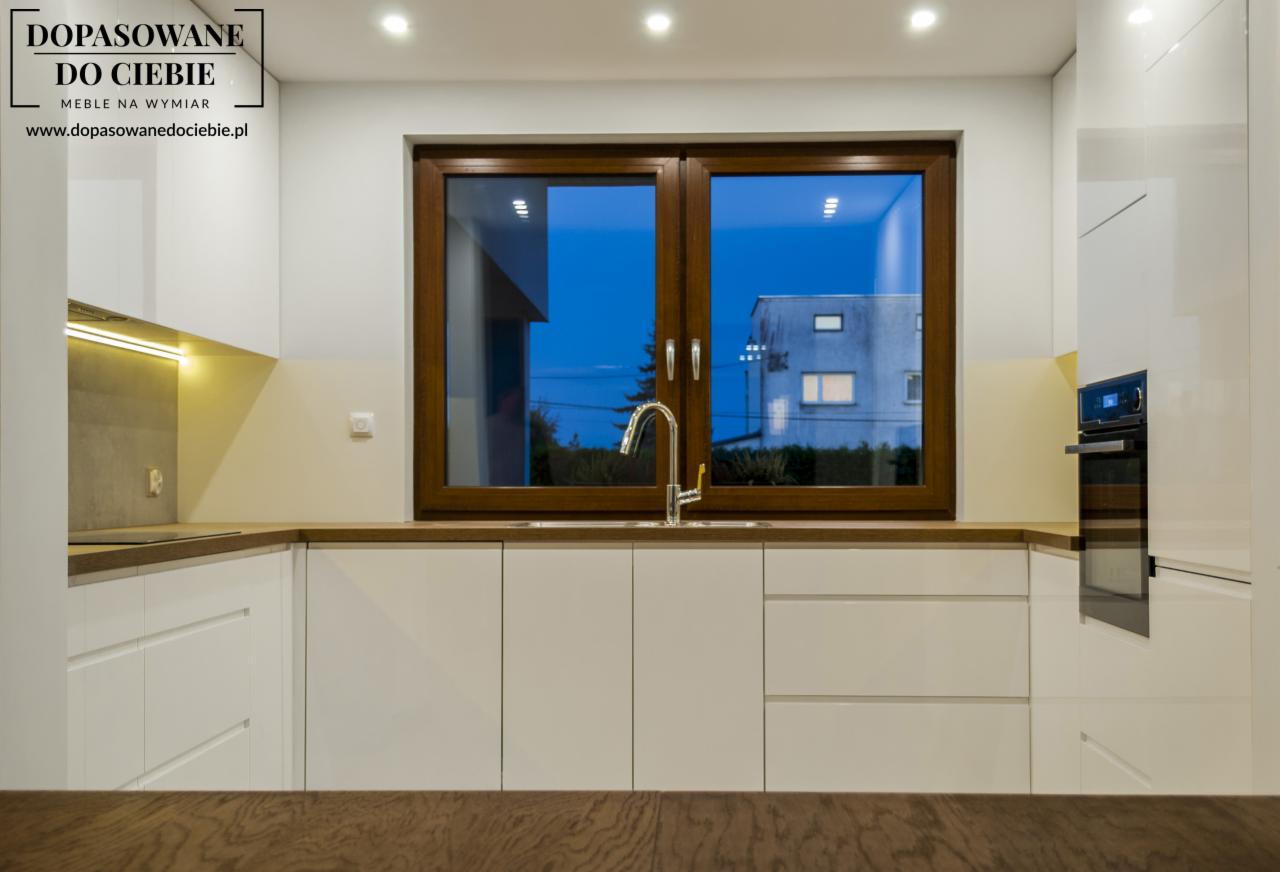 Nowoczesna, biała kuchnia z drewnianym blatem, oknem z brązowymi ramami i widokiem na oświetlony budynek w nocy.