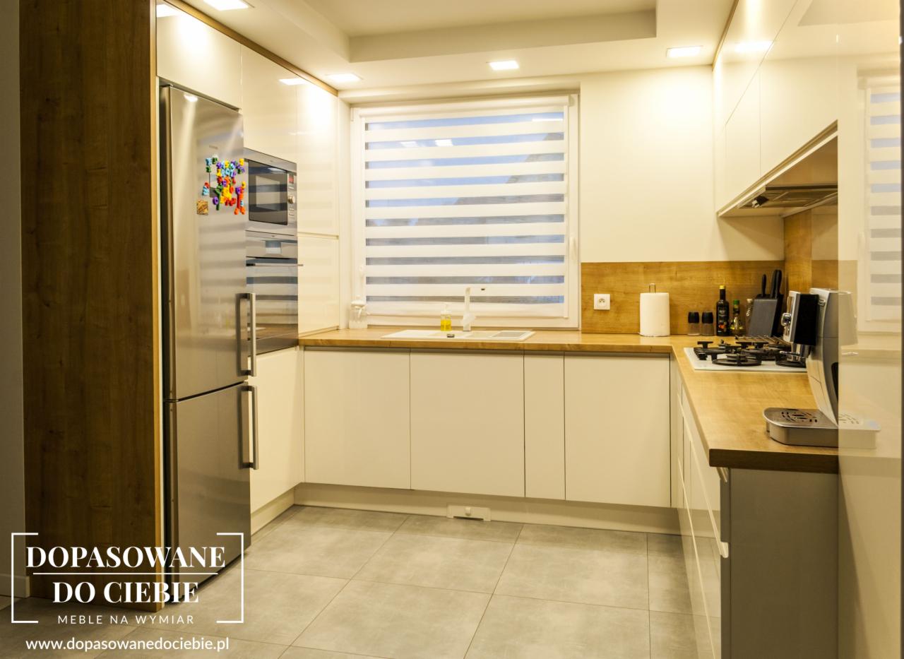 Jasna kuchnia z drewnianymi blatami, białe fronty szafek, stalowa lodówka z magnesami, okno z żaluzjami poziomymi i drewniana ściana nad blatem.