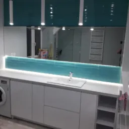 Biała zabudowa łazienkowa z umywalką, pralką i turkusową mozaiką nad blatem, oświetlona listwą LED, z dużym lustrem odbijającym fragment łazienki z grzejnikiem drabinkowym.