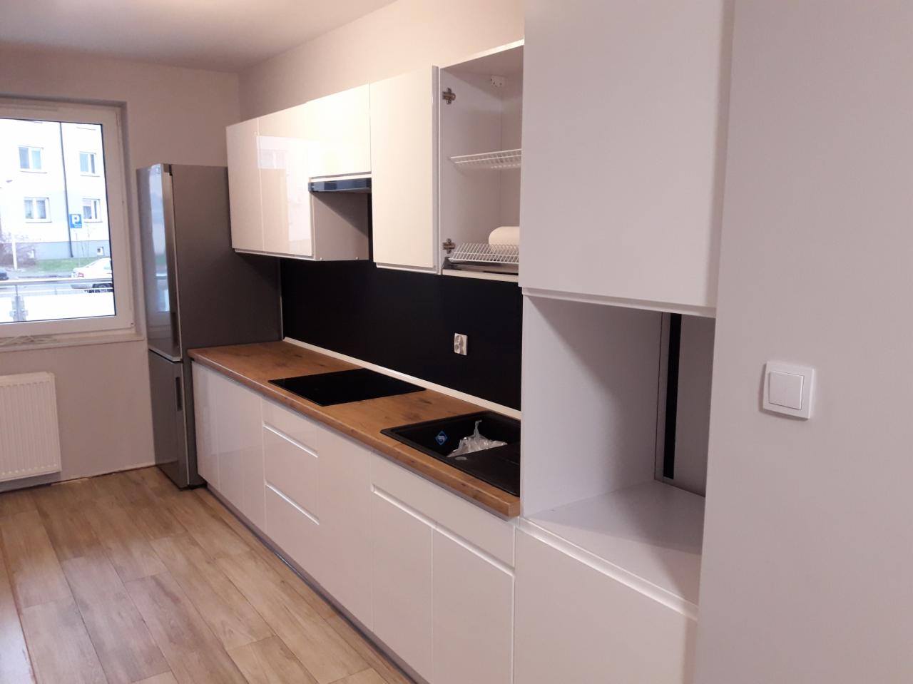 Biała, nowoczesna zabudowa kuchenna z drewnianym blatem, czarnym panelem nad blatem i otwartą szafką z metalowymi półkami, widok na okno i lodówkę.