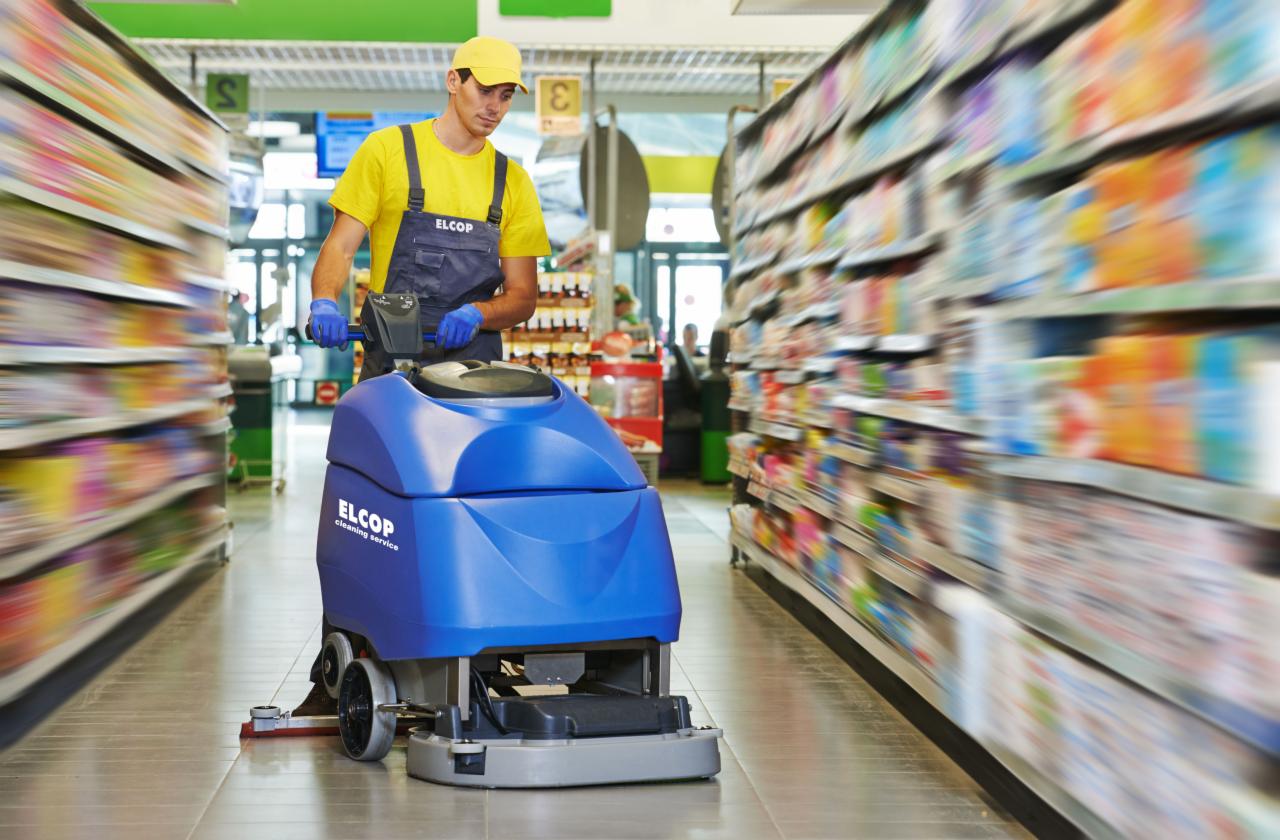 Pracownik w niebieskich rękawicach obsługuje niebieską maszynę czyszczącą podłogę w supermarkecie, półki z towarem w tle.