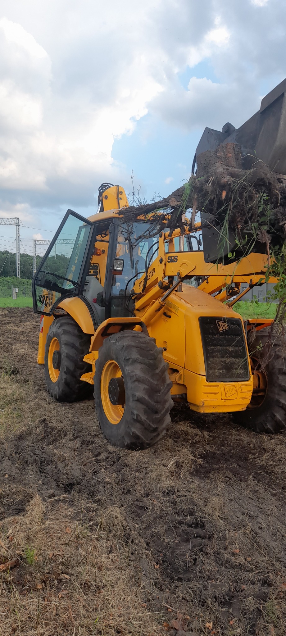 Żółta koparko-ładowarka JCB w Skawinie z otwartą kabiną i łyżką załadowaną korzeniami drzew, pracująca na polu z widocznymi liniami energetycznymi w tle.