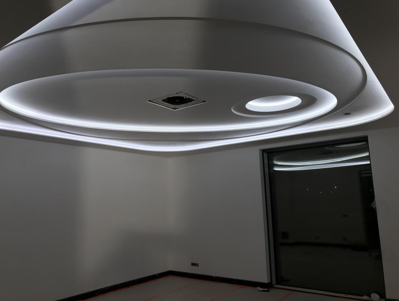 Nowoczesny sufit podwieszany z oświetleniem LED w kształcie okręgów i kwadratu, widoczny fragment ściany i okna.
