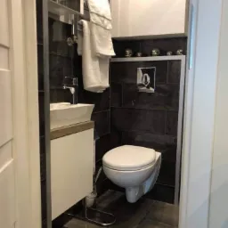 Nowoczesna łazienka z ciemnymi płytkami, wiszącą toaletą, umywalką nablatową i grzejnikiem drabinkowym z białymi ręcznikami.