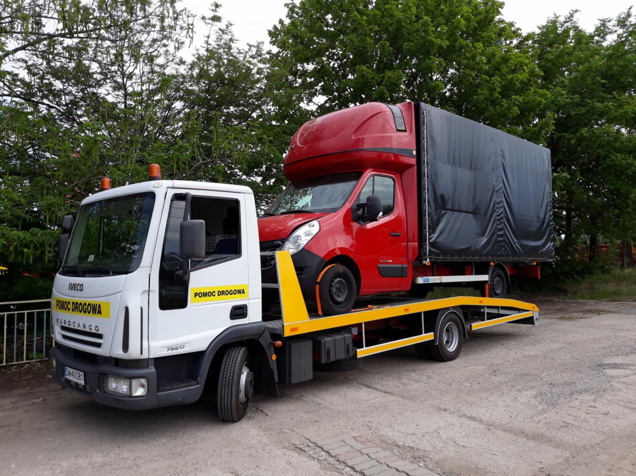 Biała ciężarówka pomocy drogowej marki Iveco z żółtymi elementami platformy transportowej przewozi czerwony samochód dostawczy z czarną plandeką na tle zielonych drzew.