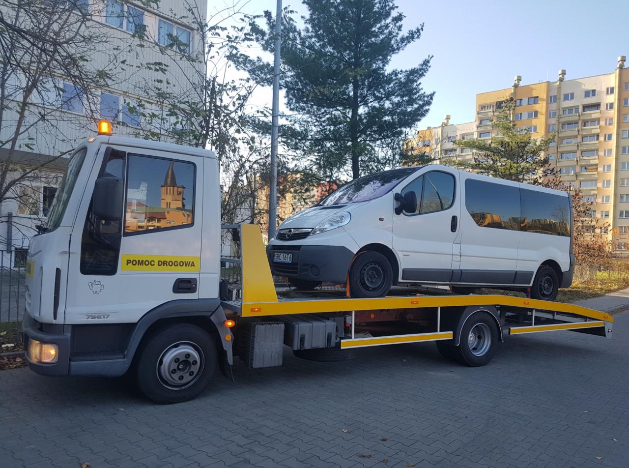 Biała pomoc drogowa przewozi białego busa na żółtej platformie w miejskiej scenerii, widoczny budynek mieszkalny w tle.