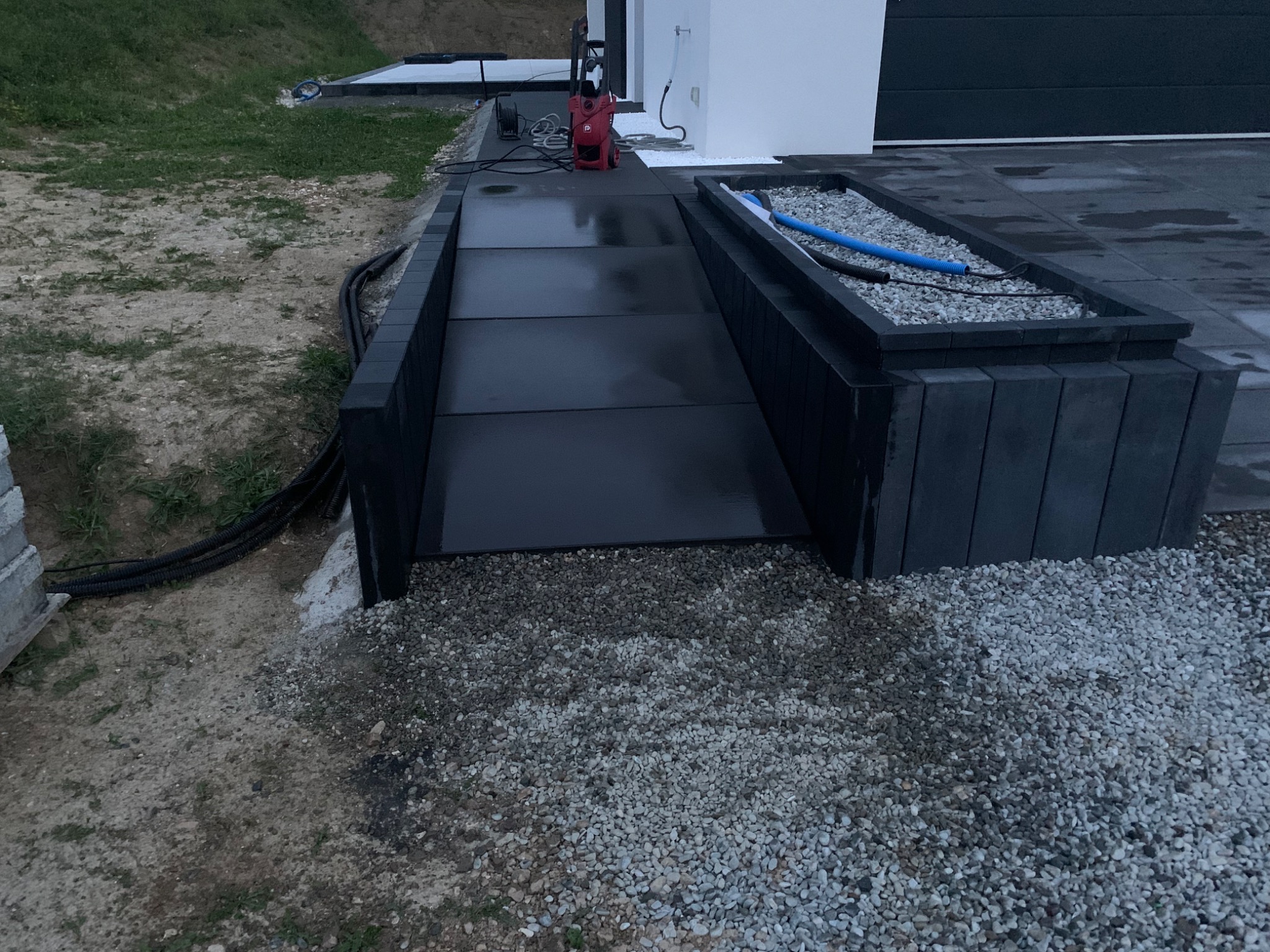 Nowoczesny podjazd z ciemnych płyt betonowych, obrzeża z prefabrykatów betonowych, rabata z białym grysem, myjka ciśnieniowa w tle.