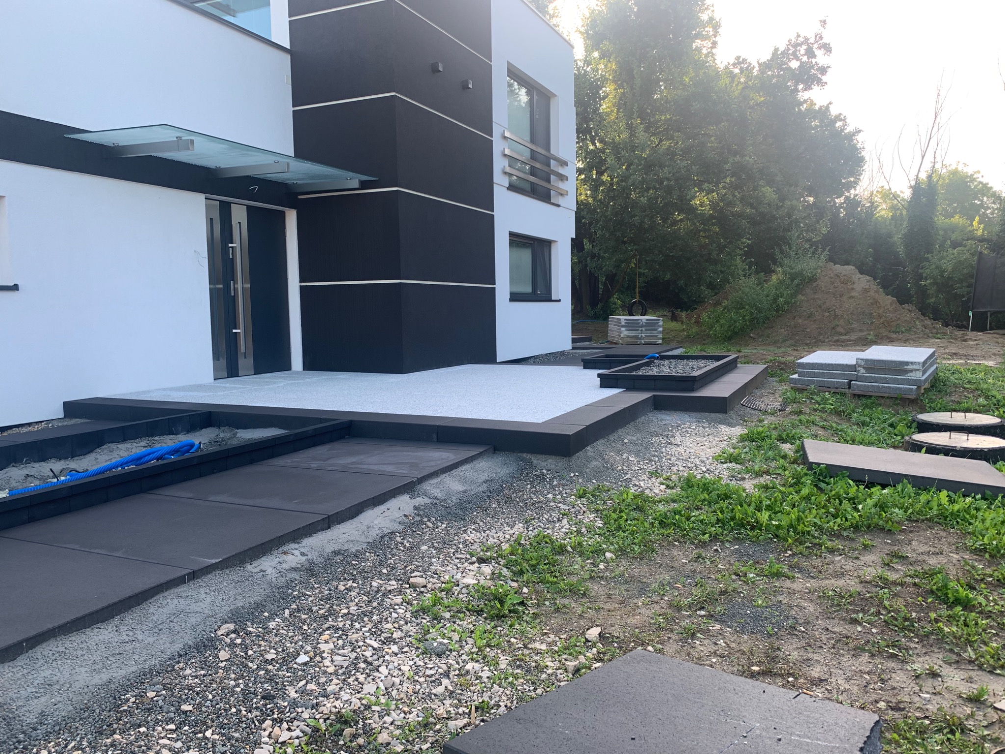 Nowoczesny dom z wykończonym wejściem z kostki brukowej, widoczny fragment ogrodu z przygotowanymi płytami do ułożenia, niebieski wąż.