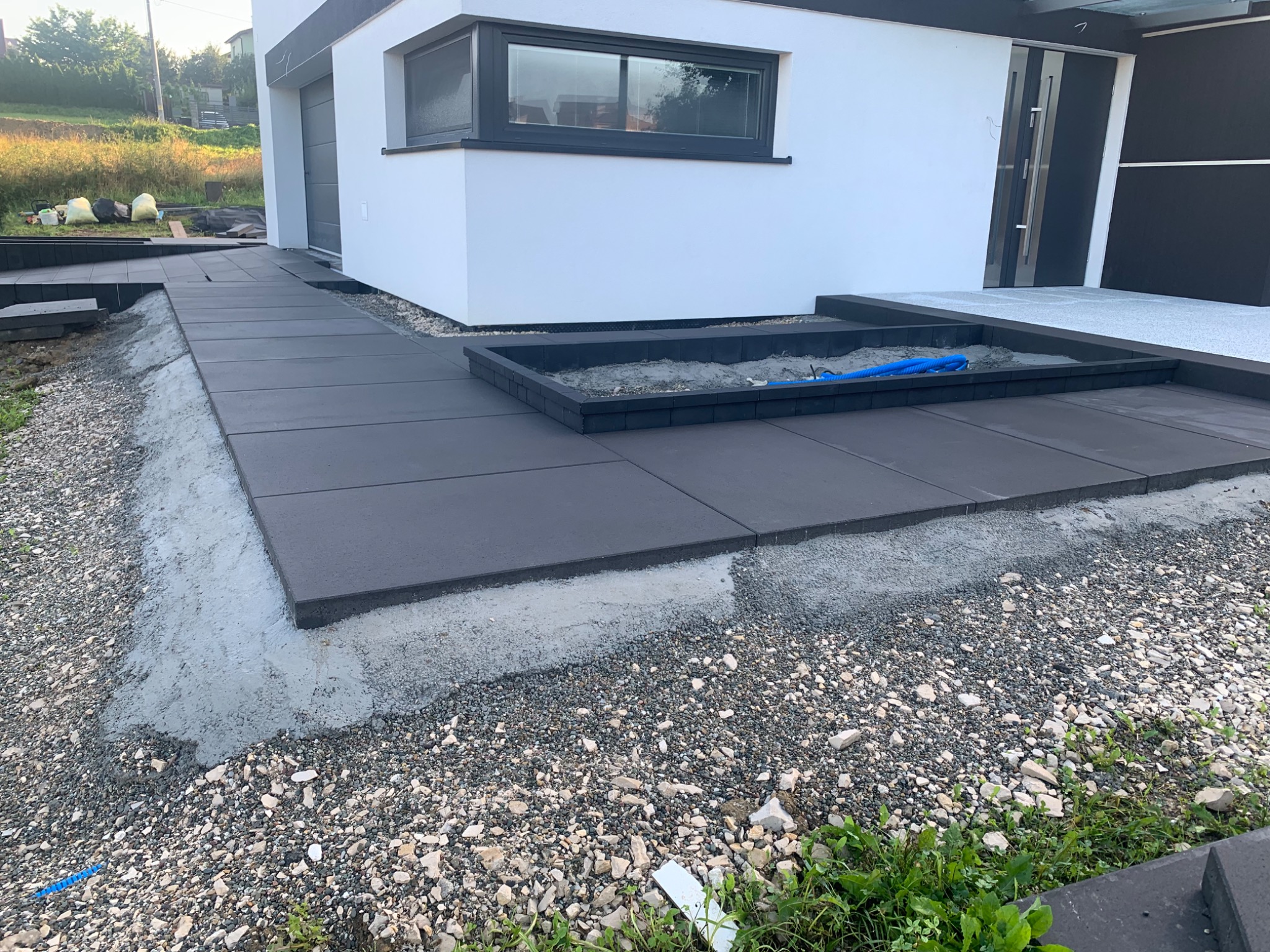 Nowoczesny podjazd z dużych, grafitowych płyt betonowych, częściowo ułożony, z widocznym podsypem i fragmentem budynku z białym tynkiem.