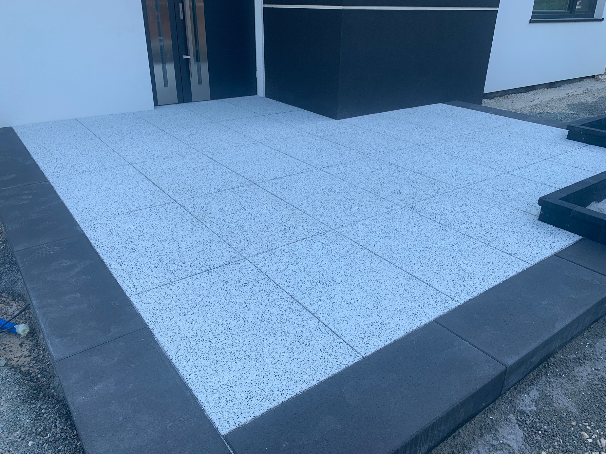 Nowoczesny taras przed domem wyłożony jasnoszarą kostką z ciemnoszarą obwódką, widoczne wejście do budynku z czarnymi elementami.