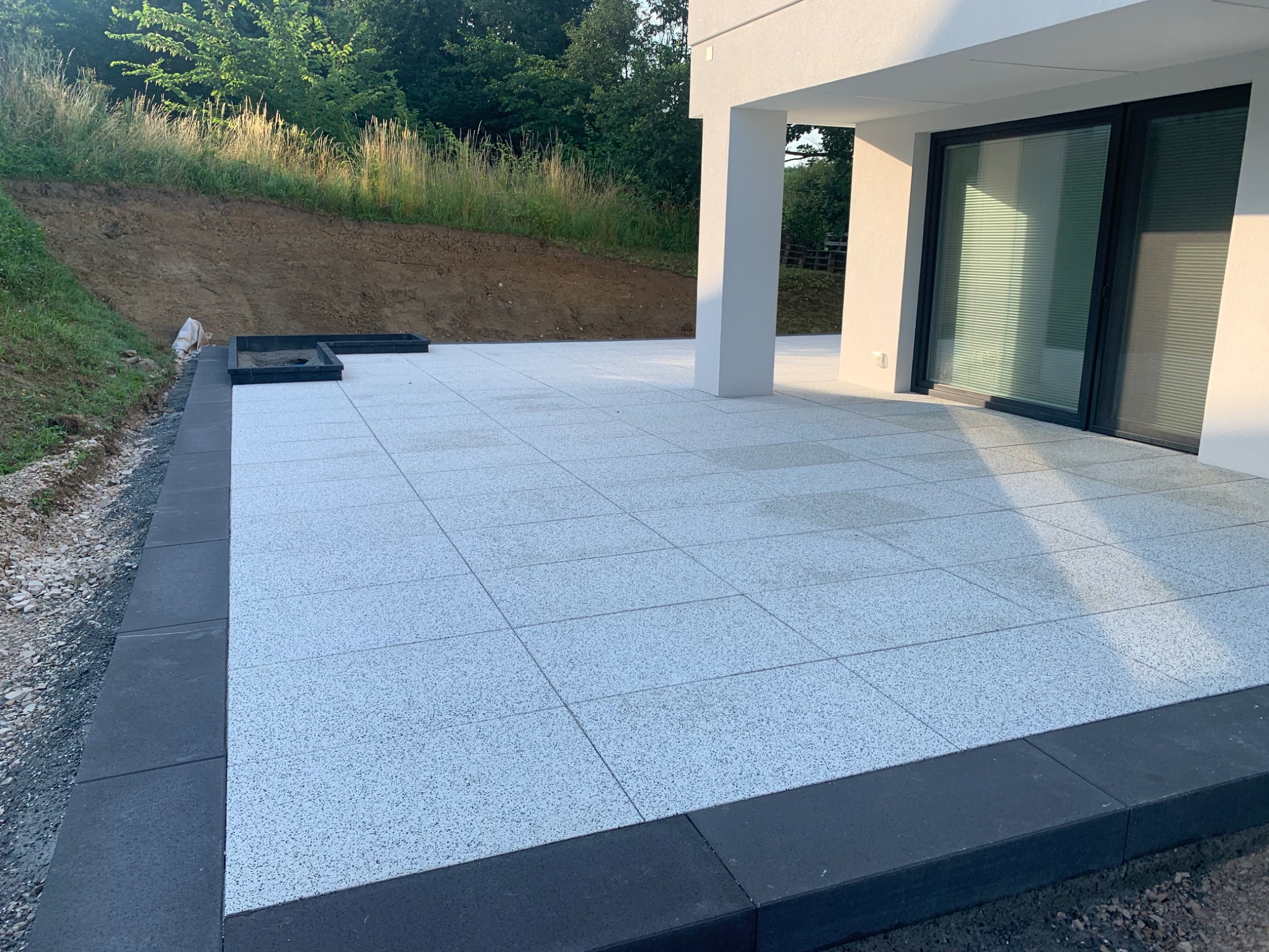 Nowoczesny taras z jasnoszarej kostki z ciemnoszarą obwódką, przylegający do białego budynku z dużymi oknami przesuwnymi, widok na zielony nasyp.