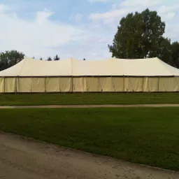 Duży, beżowy namiot imprezowy rozstawiony na trawiastym terenie, widoczne drzewa w tle i fragment nieba.