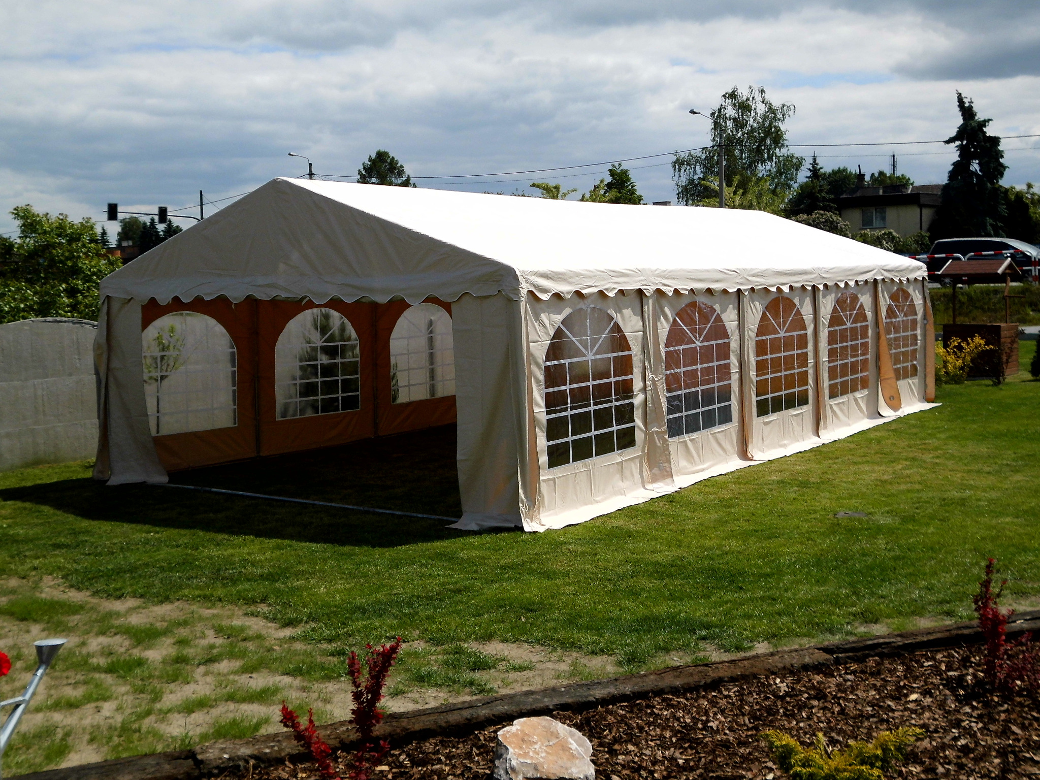 Beżowy namiot imprezowy z oknami na trawniku, widok z zewnątrz, w tle domy i sygnalizacja świetlna.