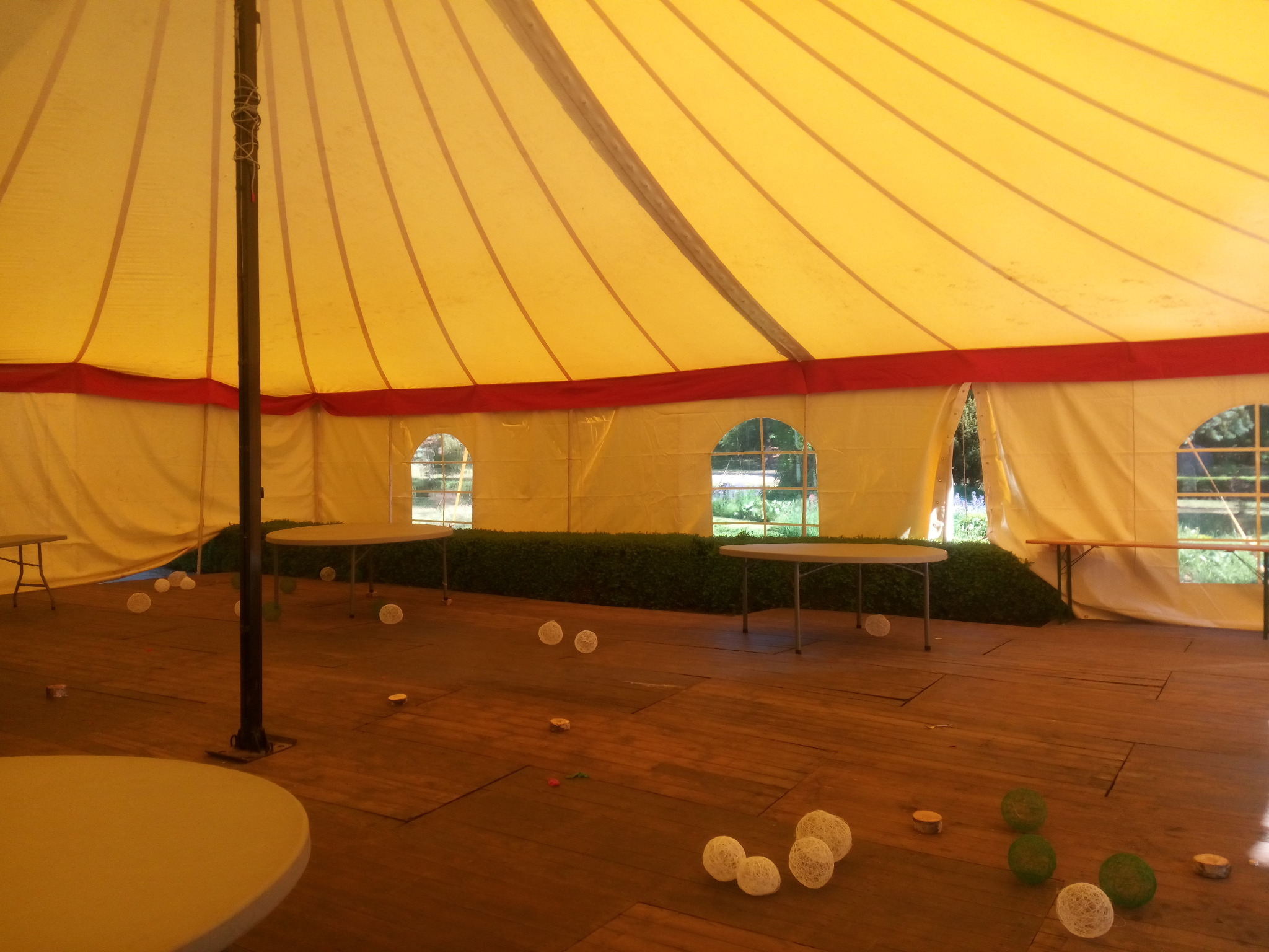 Wnętrze żółtego namiotu z drewnianą podłogą, okrągłymi stołami i dekoracjami w postaci białych i zielonych kul z włóczki. Widoczne okrągłe okna z widokiem na zieleń na zewnątrz.