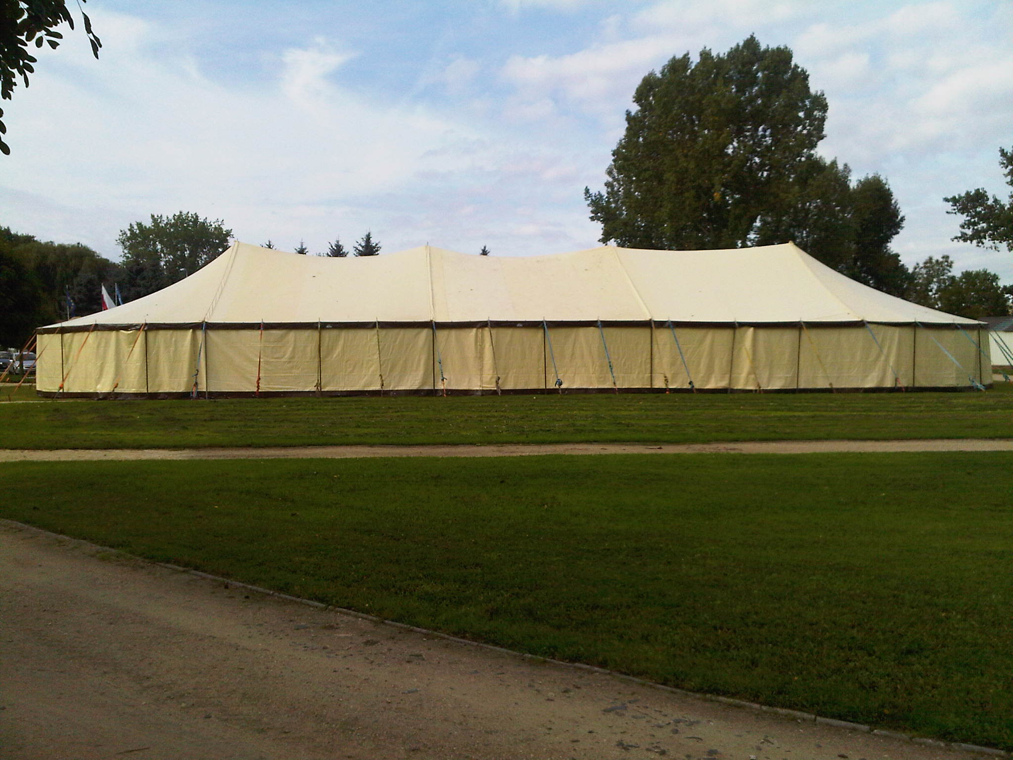 Duży, beżowy namiot imprezowy rozstawiony na trawiastym terenie, widoczne drzewa w tle i fragment nieba.