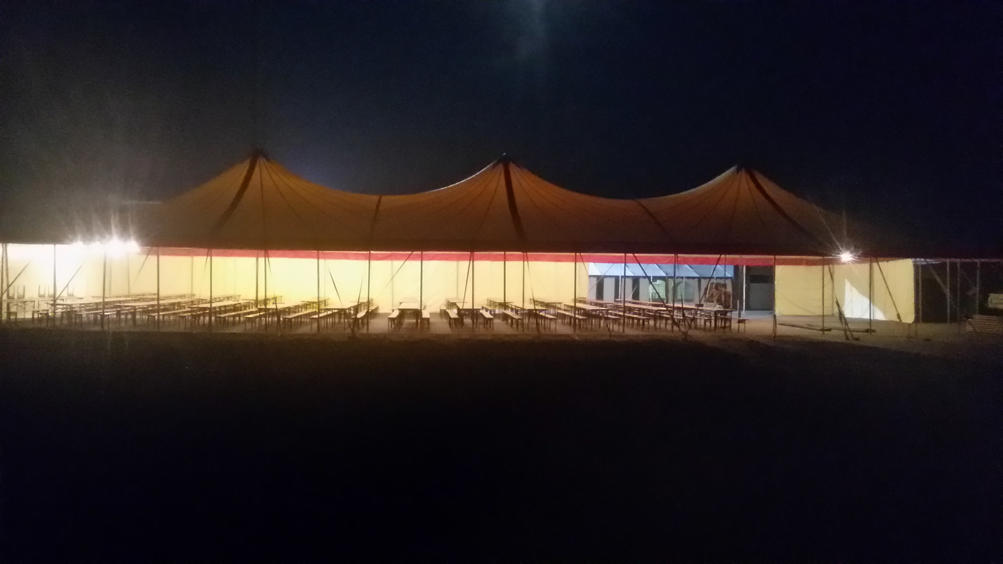 Oświetlony namiot imprezowy z rzędami drewnianych stołów i ławek w nocy.