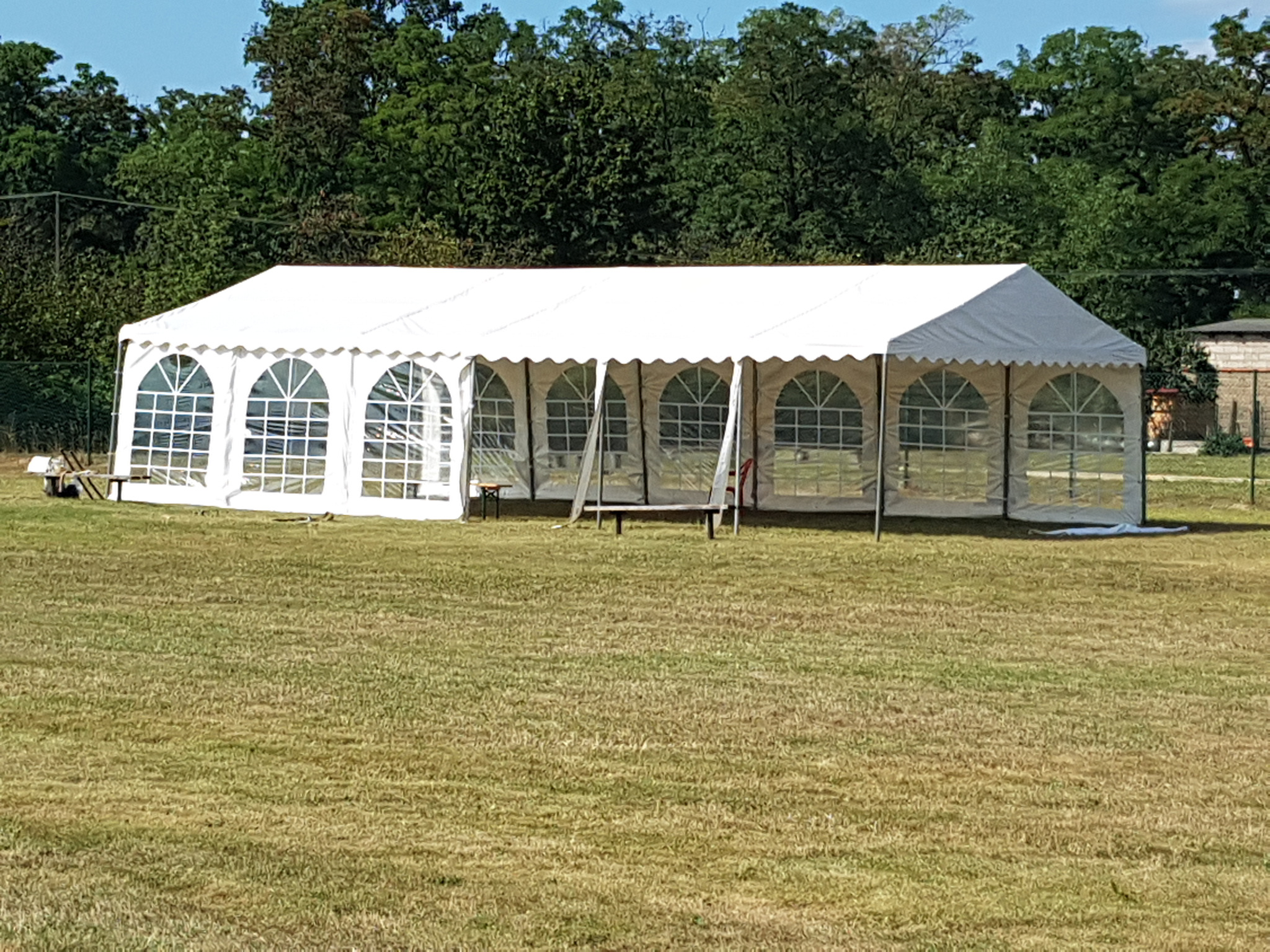 Biały namiot plenerowy z oknami na trawniku, widok z przodu, lekko z prawej strony, drzewa w tle.