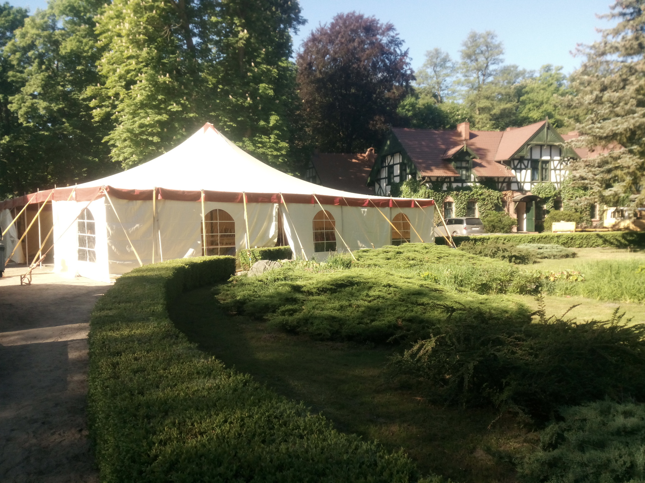 Duży, biały namiot eventowy z oknami w ogrodzie z ozdobnymi krzewami i budynkiem w stylu rustykalnym w tle.