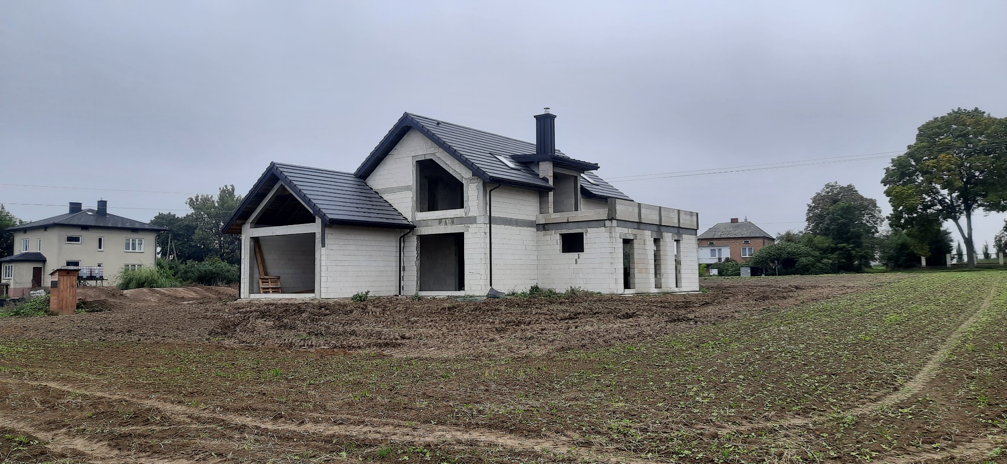 Niedokończony budynek z białych pustaków z czarnym dachem na tle zaoranego pola i dwóch innych domów.