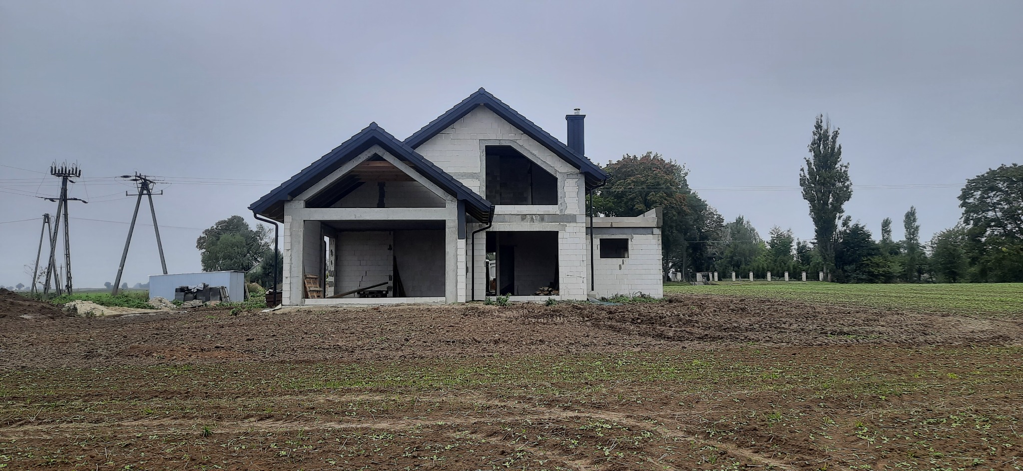 Surowy stan domu z białego bloczka, widoczny komin, czarne rynny i częściowo wykonany dach na tle pola i słupów energetycznych