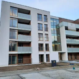 Nowoczesny budynek apartamentowy z białymi elewacjami, szklanymi balkonami i drewnianymi akcentami, widoczny z poziomu brukowanego placu z elementami placu budowy.