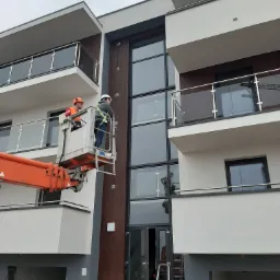 Dwóch pracowników w podnośniku koszowym na tle nowoczesnego budynku wielorodzinnego z dużymi przeszkleniami, prawdopodobnie podczas prac konserwacyjnych lub inspekcyjnych.
