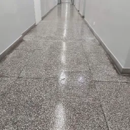 Długi korytarz z błyszczącą, umytą podłogą z lastryko, widoczne odbicia światła, szare ściany z białymi drzwiami po lewej stronie i widok na dalszą część korytarza z drzwiami na końcu.