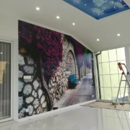 Pokój z fototapetą przedstawiającą kamienną uliczkę z kwiatami i niebieską ławką, podświetlany sufit imitujący niebo z gwiazdami, białe płytki na podłodze, drabina oparta o okno.