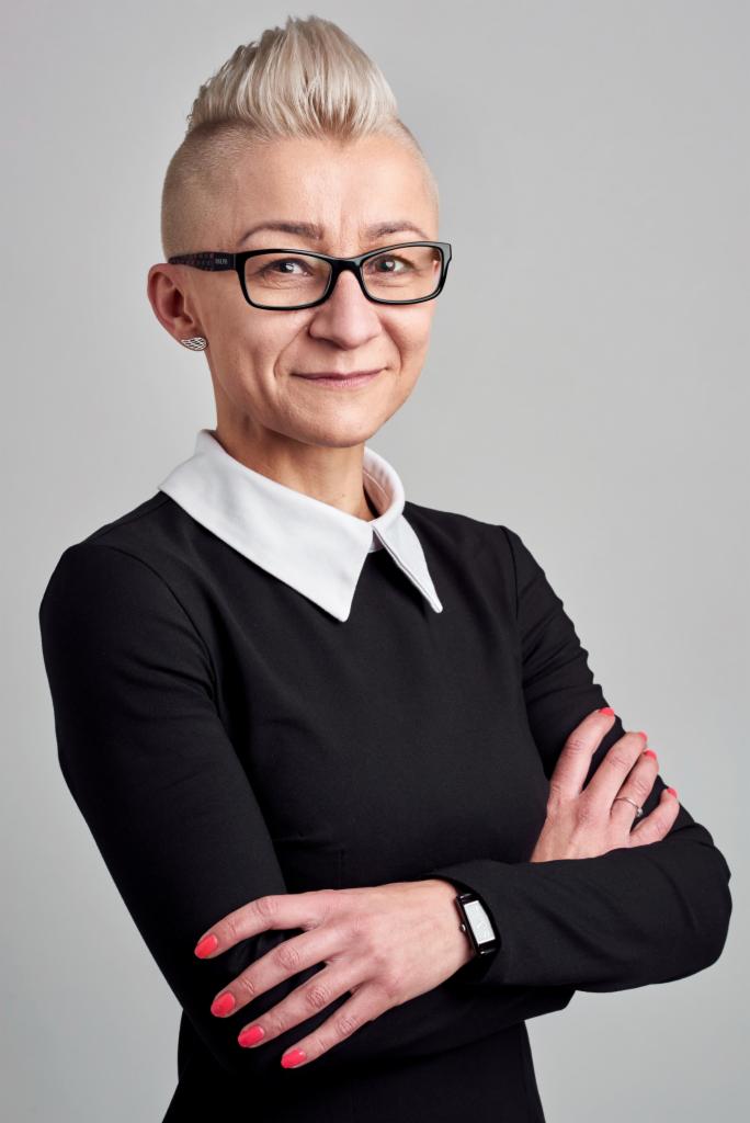 Portret kobiety w czarnej sukience z białym kołnierzykiem i okularach, z rękami skrzyżowanymi na piersiach, na jednolitym szarym tle.