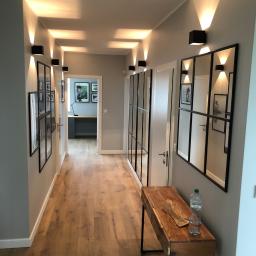 Sym-Bud Piotr Symon - Nowoczesny korytarz z szarą ścianą, drewnianą podłogą, czarnymi kinkietami oświetlającymi sufit, dekoracyjnymi lustrami w czarnych ramach, galerią zdjęć i drewnianą konsolą z butelką wody.