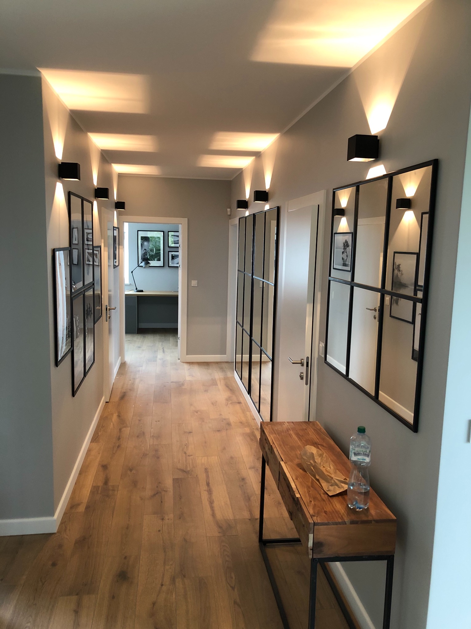 Nowoczesny korytarz z szarą ścianą, drewnianą podłogą, czarnymi kinkietami oświetlającymi sufit, dekoracyjnymi lustrami w czarnych ramach, galerią zdjęć i drewnianą konsolą z butelką wody.