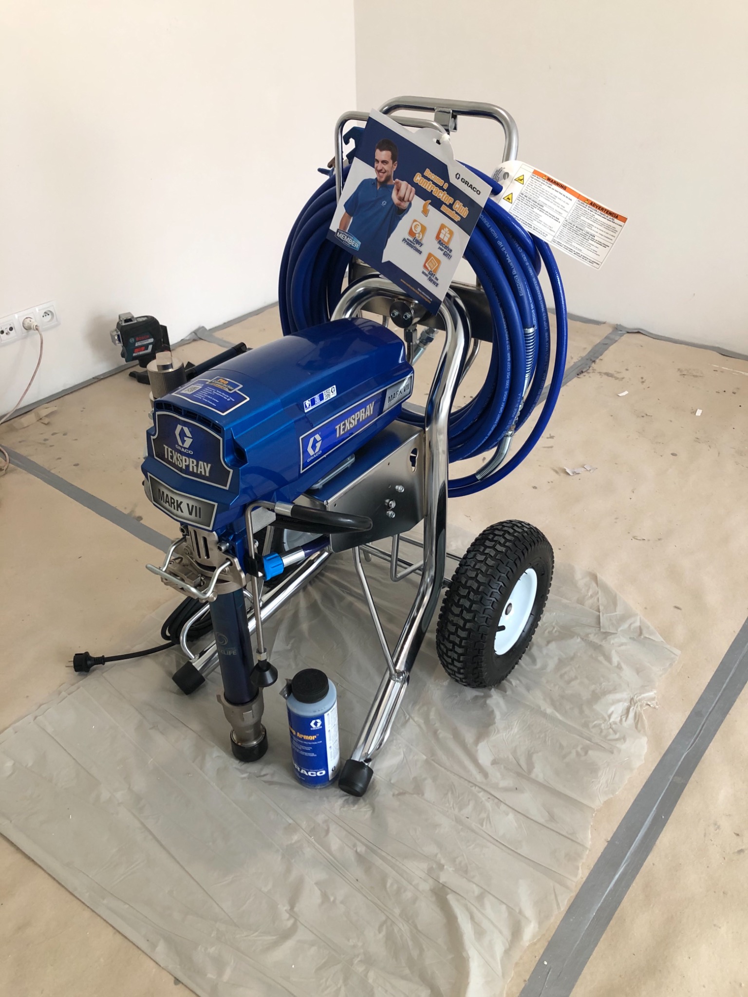 Niebieski agregat malarski TexSpray Mark VII na kółkach, z rozwiniętym wężem, stojący na zabezpieczonej folią podłodze w trakcie przygotowań do malowania ścian. W tle widoczny laser krzyżowy.