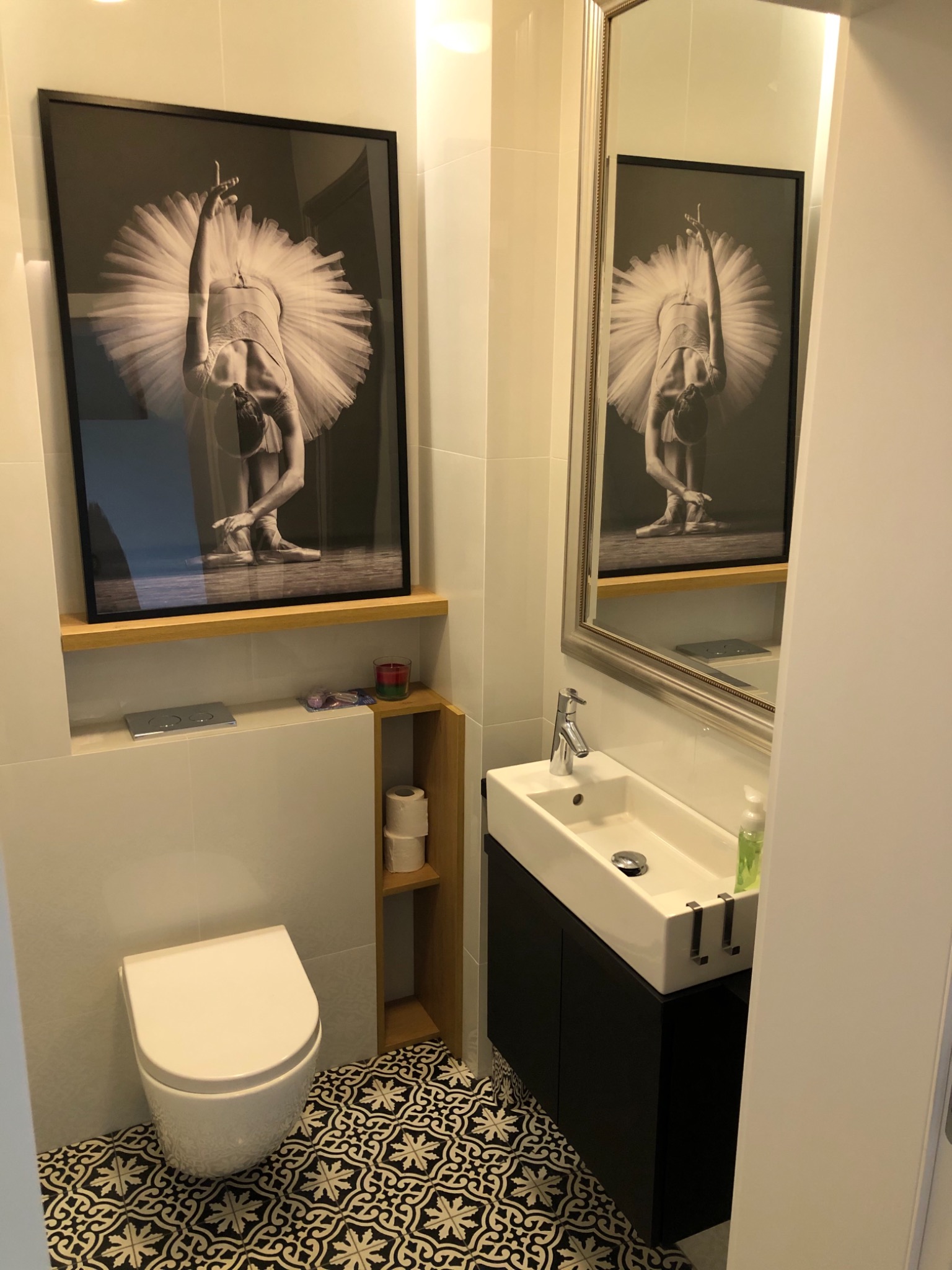 Nowoczesna łazienka z białymi płytkami, czarno-białą podłogą w geometryczne wzory, wiszącą toaletą, umywalką na szafce i czarno-białym zdjęciem baletnicy w tutu w ramie.